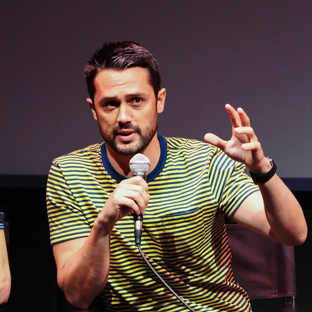 Stephen Colletti speaking in Talking Chernoby holding a microphone wearing a yellow t-shirt with blue stripes on it.